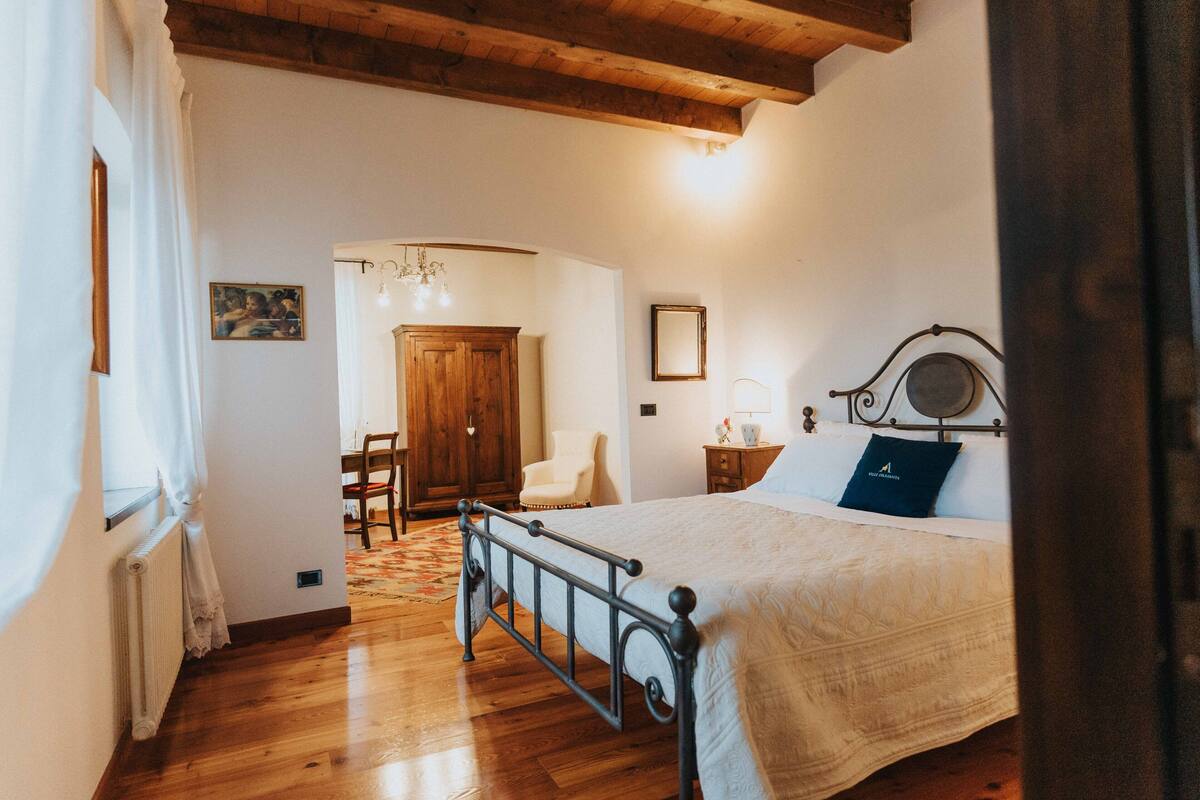 Un dormitorio con suelo de madera, una cama con estructura de metal, una silla, un armario de madera y una mesita de noche con lámpara.