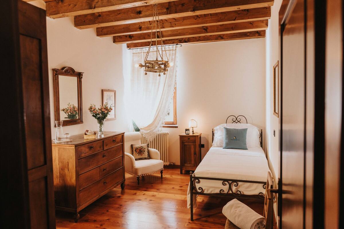 Un dormitorio con piso de madera, una cama con estructura metálica, un aparador de madera, una silla, una mesita de noche con lámpara y una ventana con cortinas translúcidas.