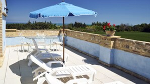 Un patio con sillones blancos, una sombrilla azul y vista al mar.