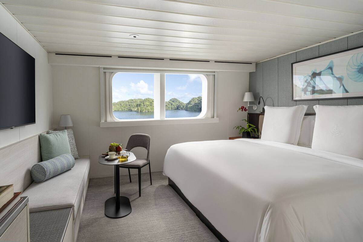 A hotel room with a large bed, a small table with fruit, a chair, a TV, and a window with a view of a lake.