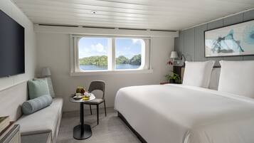 Habitación de hotel con una cama grande, una mesita con fruta, una silla, un televisor y una ventana con vista a un lago.