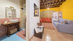 Un baño con lavamanos, espejo y ducha. Hay un sofá y una mesa con un libro y un florero en la sala.