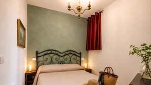 Ein Schlafzimmer mit einem Bett, Nachttischlampen, einem Wandbild, einem roten Vorhang, einem Korbwägelchen und einer Vase mit Pflanze.