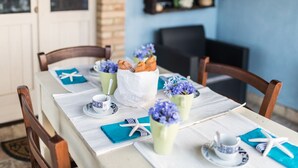 Ein Esstisch ist gedeckt mit einer weißen Tischdecke, blauen Servietten und einem weißen Serviettenhalter. Es sind zwei weiße Tassen mit blauen Mustern, eine grüne Tasse mit einem Löffel und eine weiße Papiertüte mit Gebäck zu sehen.