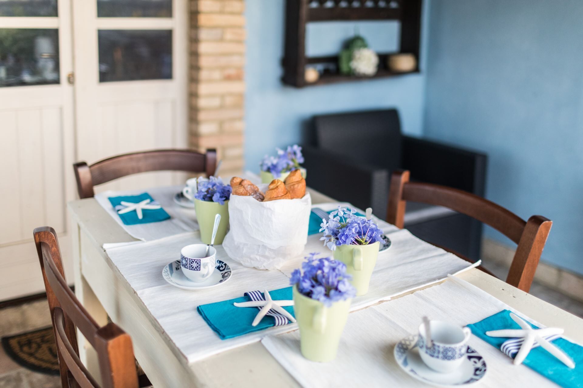 Ein Esstisch ist gedeckt mit einer weißen Tischdecke, blauen Servietten und einem weißen Serviettenhalter. Es sind zwei weiße Tassen mit blauen Mustern, eine grüne Tasse mit einem Löffel und eine weiße Papiertüte mit Gebäck zu sehen.