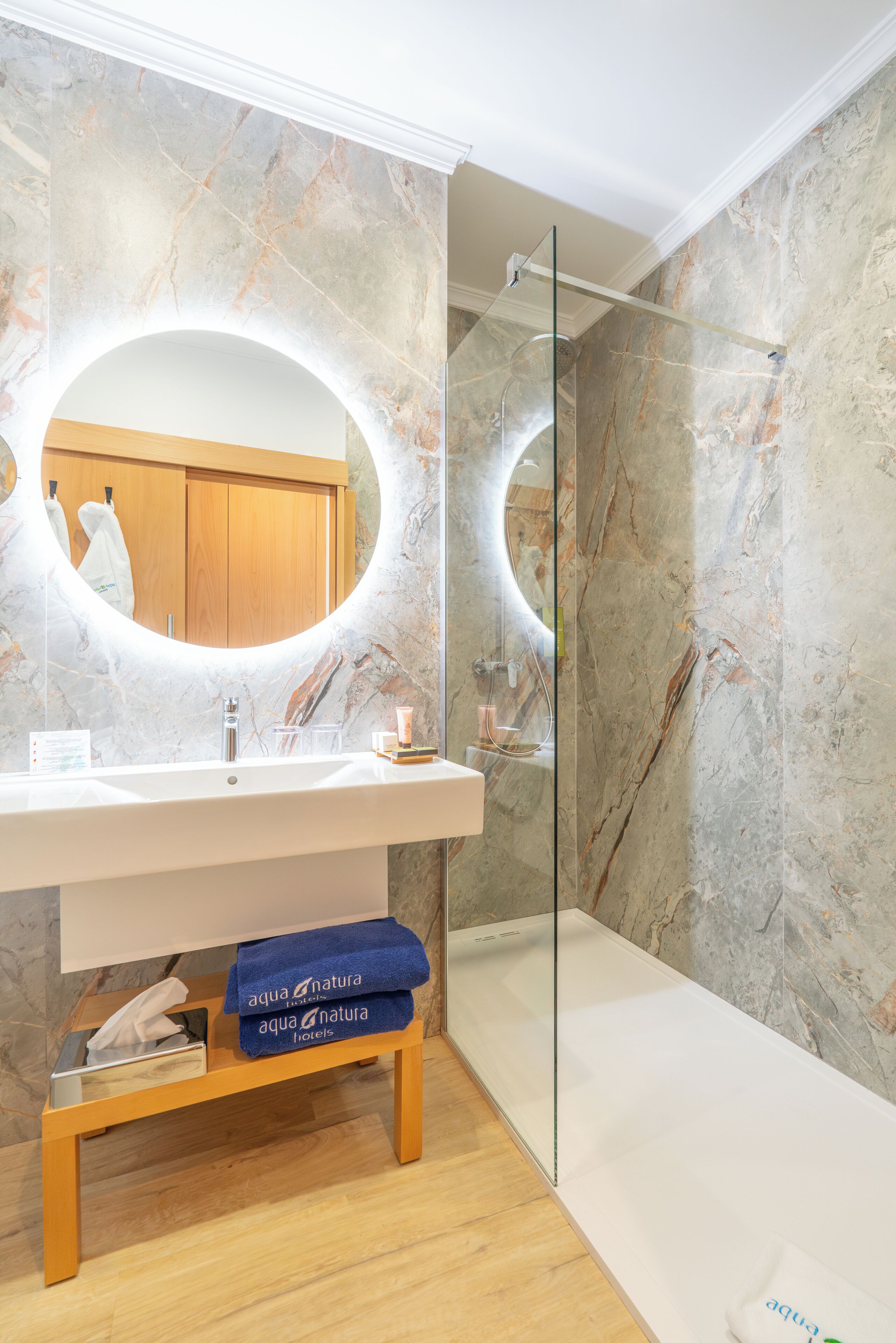 A modern bathroom with a marble wall, a large oval mirror, a glass-enclosed shower, and a wooden bench with towels.