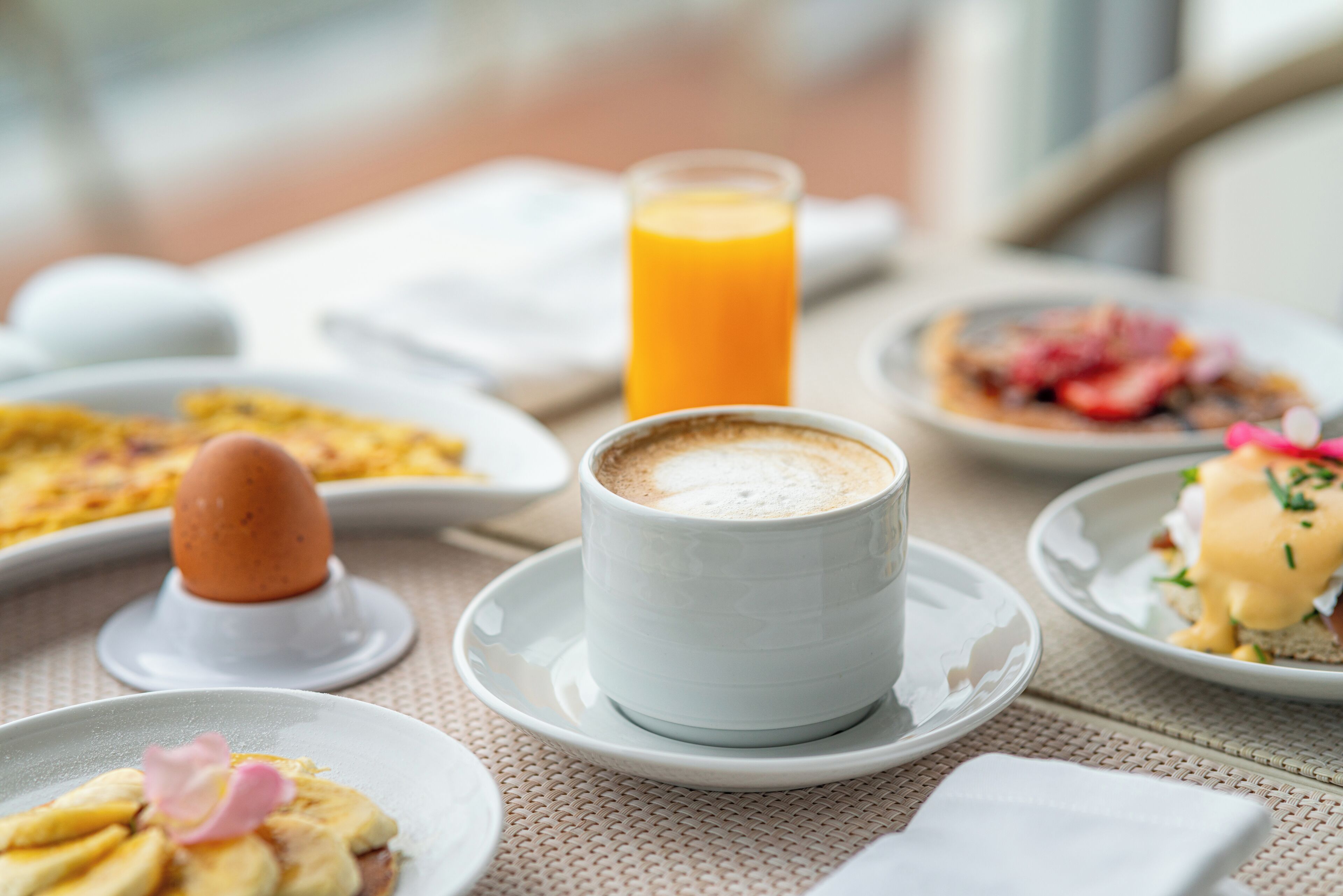 A breakfast setting with a cup of coffee, a glass of orange juice, and various dishes including eggs, pancakes, and a dessert with a pink flower garnish.