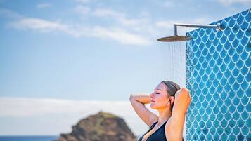 A woman in a black swimsuit under a shower with a blue tiled wall and a rocky landscape in the background.