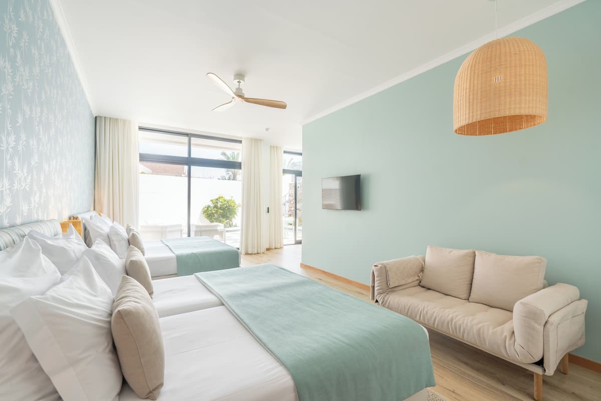 A hotel room with two beds, a sofa, a ceiling fan, a TV, and a large window with curtains.