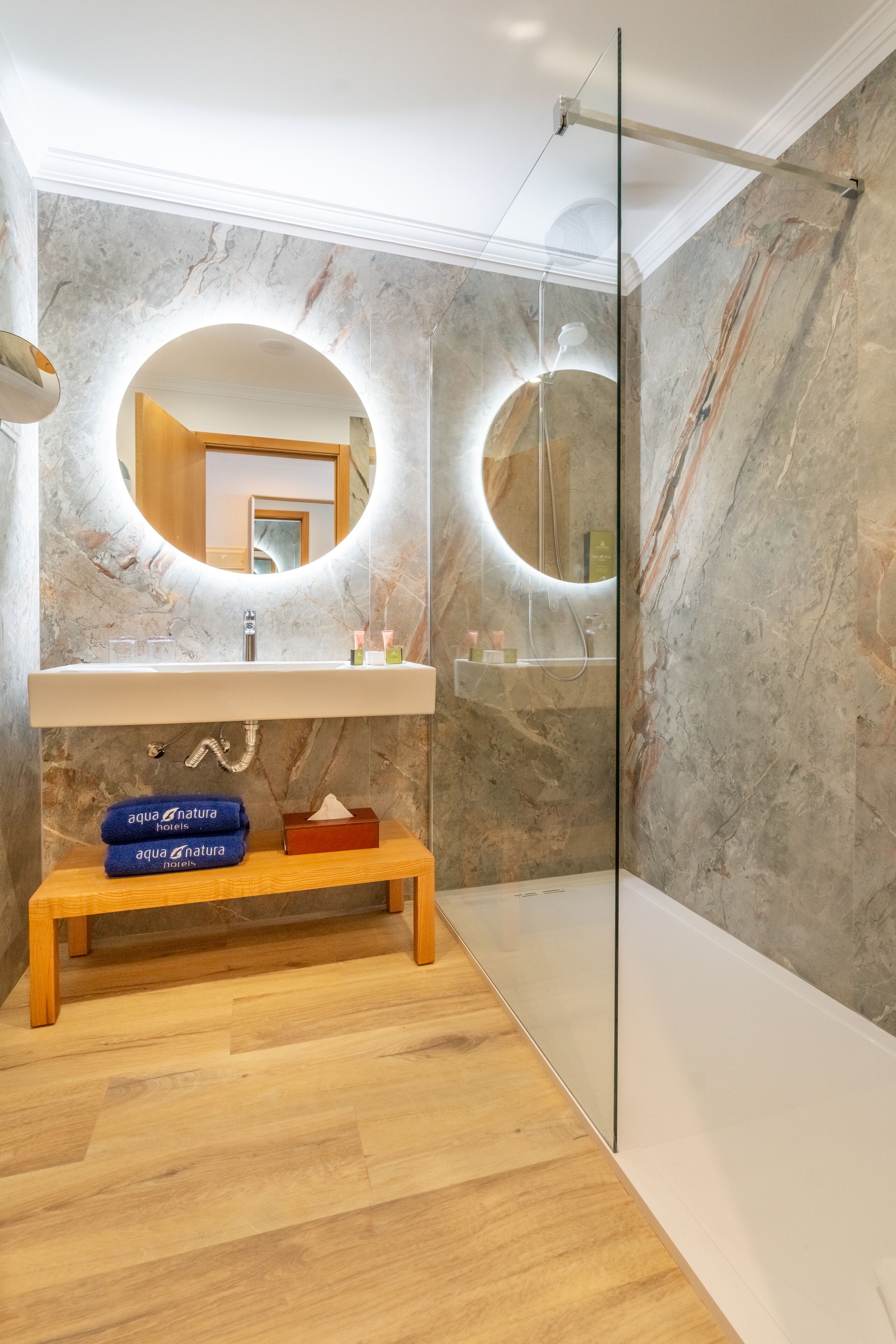 A modern bathroom with a marble wall, a wooden bench, and a glass shower enclosure.