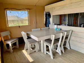 Une table à manger en bois, accompagnée de six chaises, une fenêtre donnant sur un paysage verdoyant et un mur en bois sur lequel est fixé un porte-manteau.