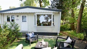 A small, white, single-story building with a dark roof, surrounded by lush greenery and trees. - Cosy cottage at Veluwe (Putten)