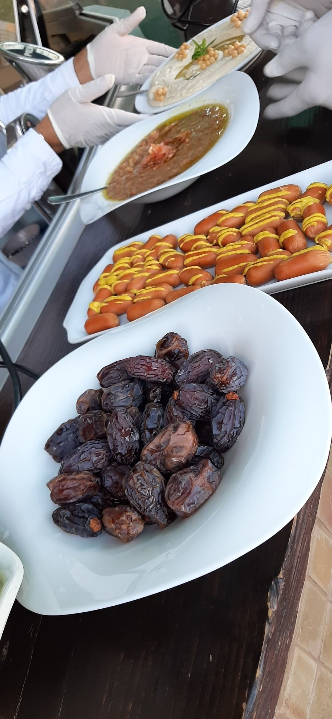 Una mesa de buffet con diversos platos, incluyendo un tazón de dátiles, un plato de perros calientes y un recipiente con salsa verde.