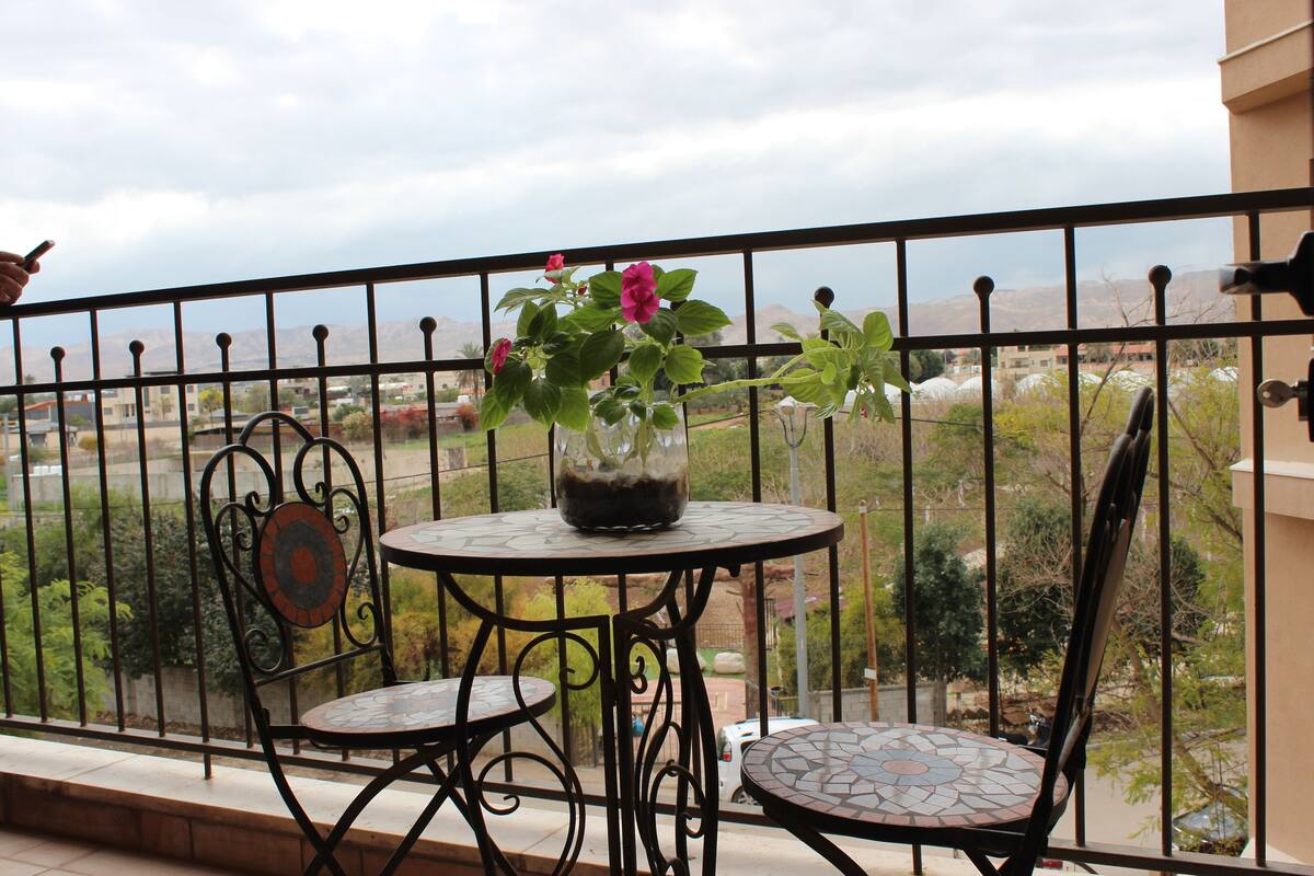 Ein Balkon mit rundem Tisch und zwei Stühlen, ein Blumentopf mit Blumen sowie ein Blick auf eine gebirgige Landschaft.