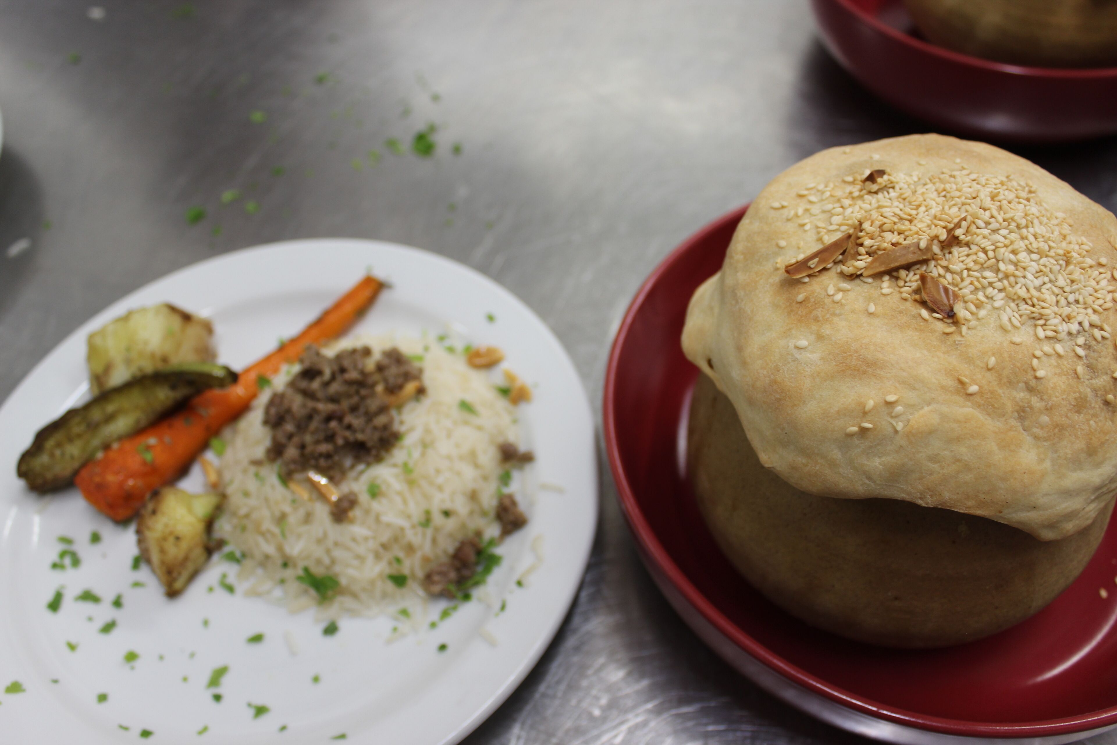 A plate with rice, vegetables, and a meat dish, and a red bowl with a bread roll.