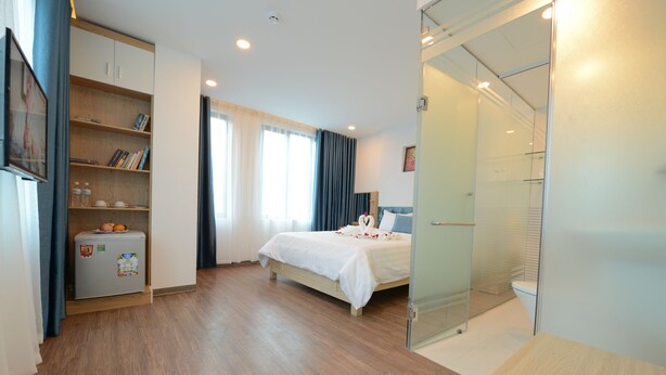 A modern hotel room with a wooden floor, a bed with white bedding, a small refrigerator, a bookshelf, and a glass shower enclosure. - Hanoi Prime Center Hotel & Spa (Hanoi)