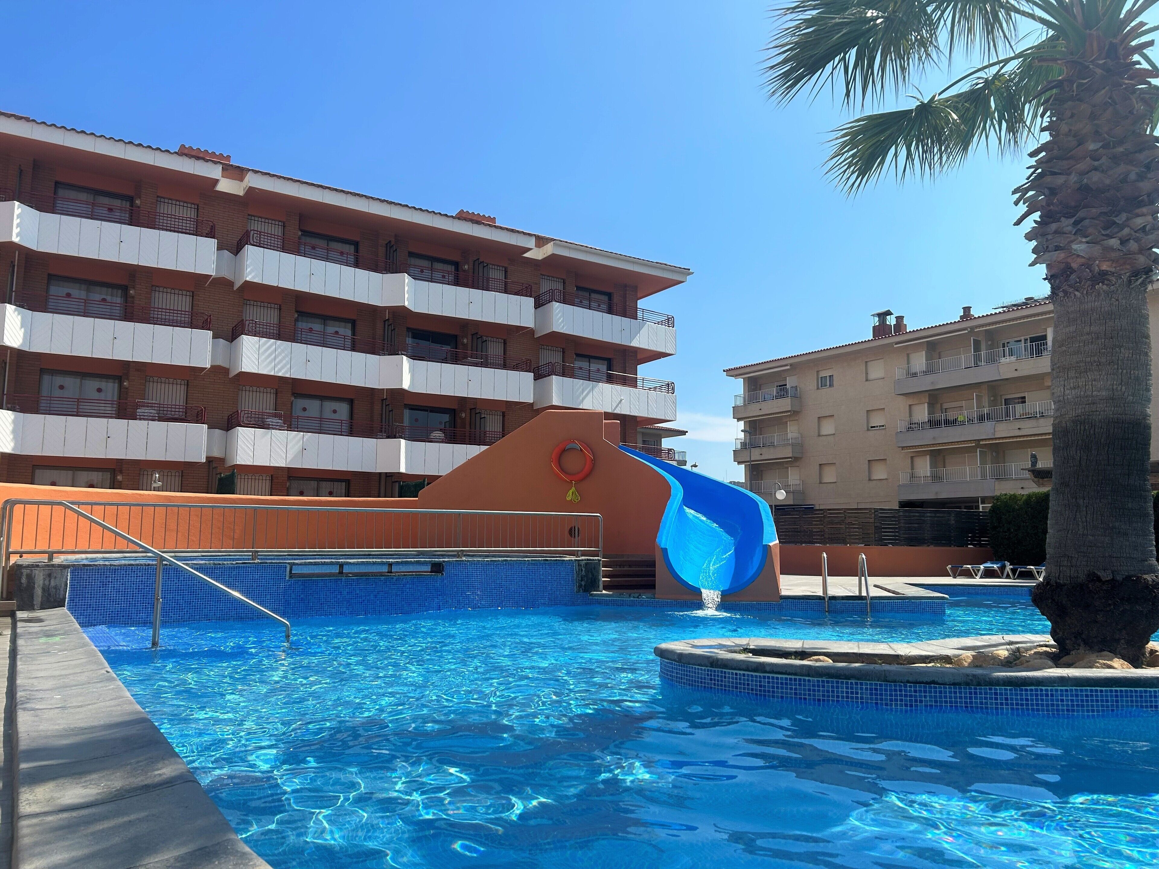 A swimming pool with a water slide, surrounded by multi-story buildings with balconies.