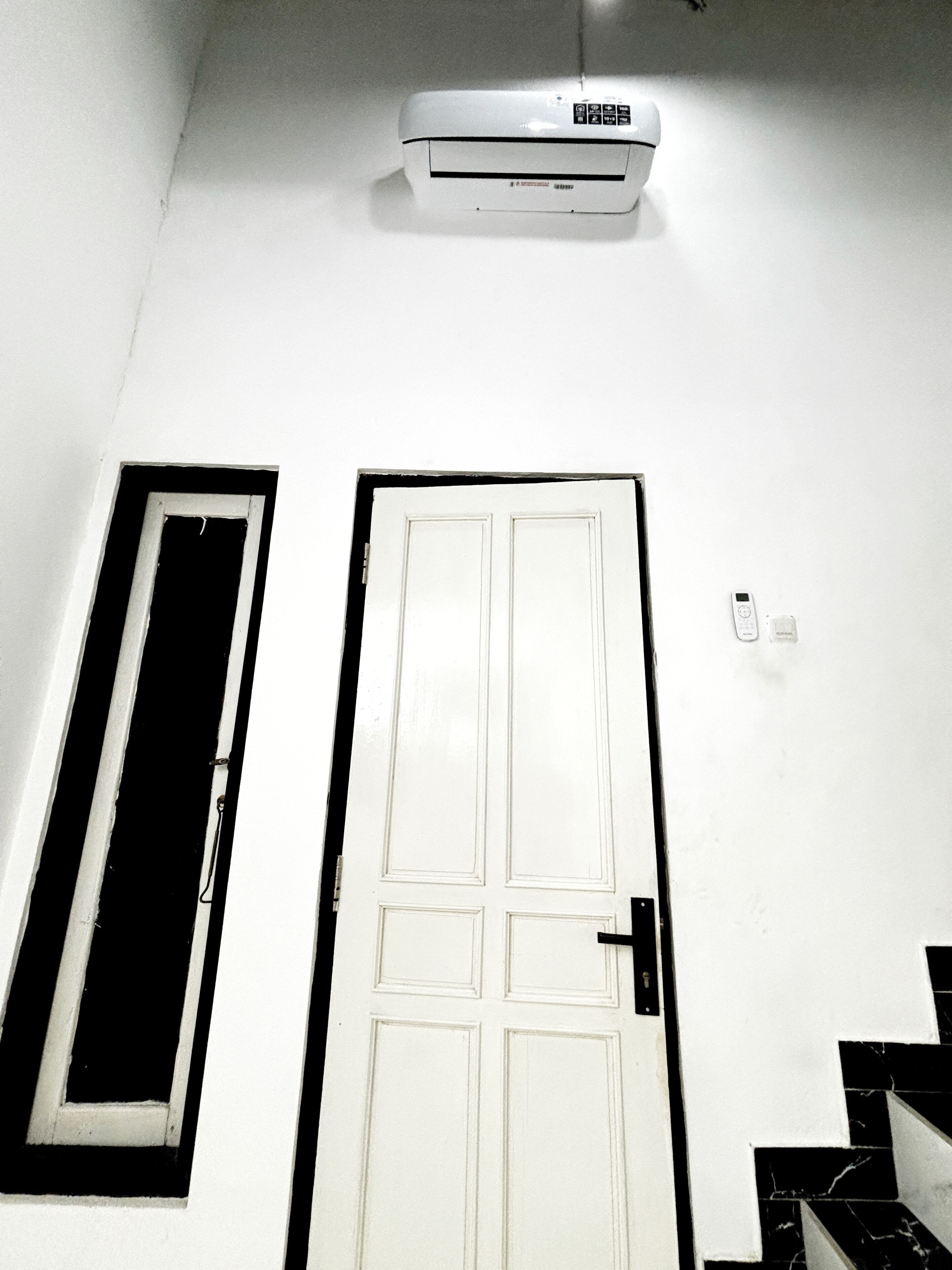 A white door with a window to the left, a wall-mounted air conditioner above, and a small electrical outlet to the right.