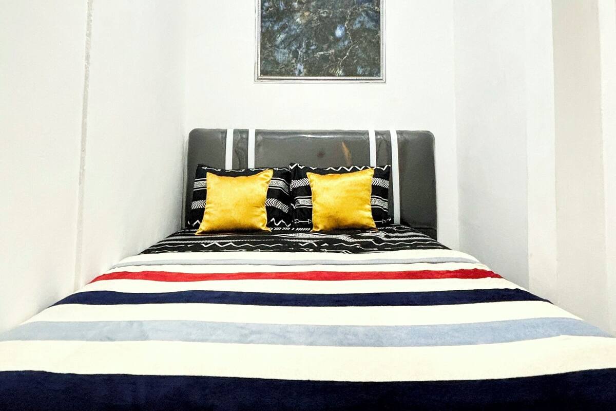 A small, narrow room with a bed, a wall-mounted air conditioner, and a framed picture above the bed.