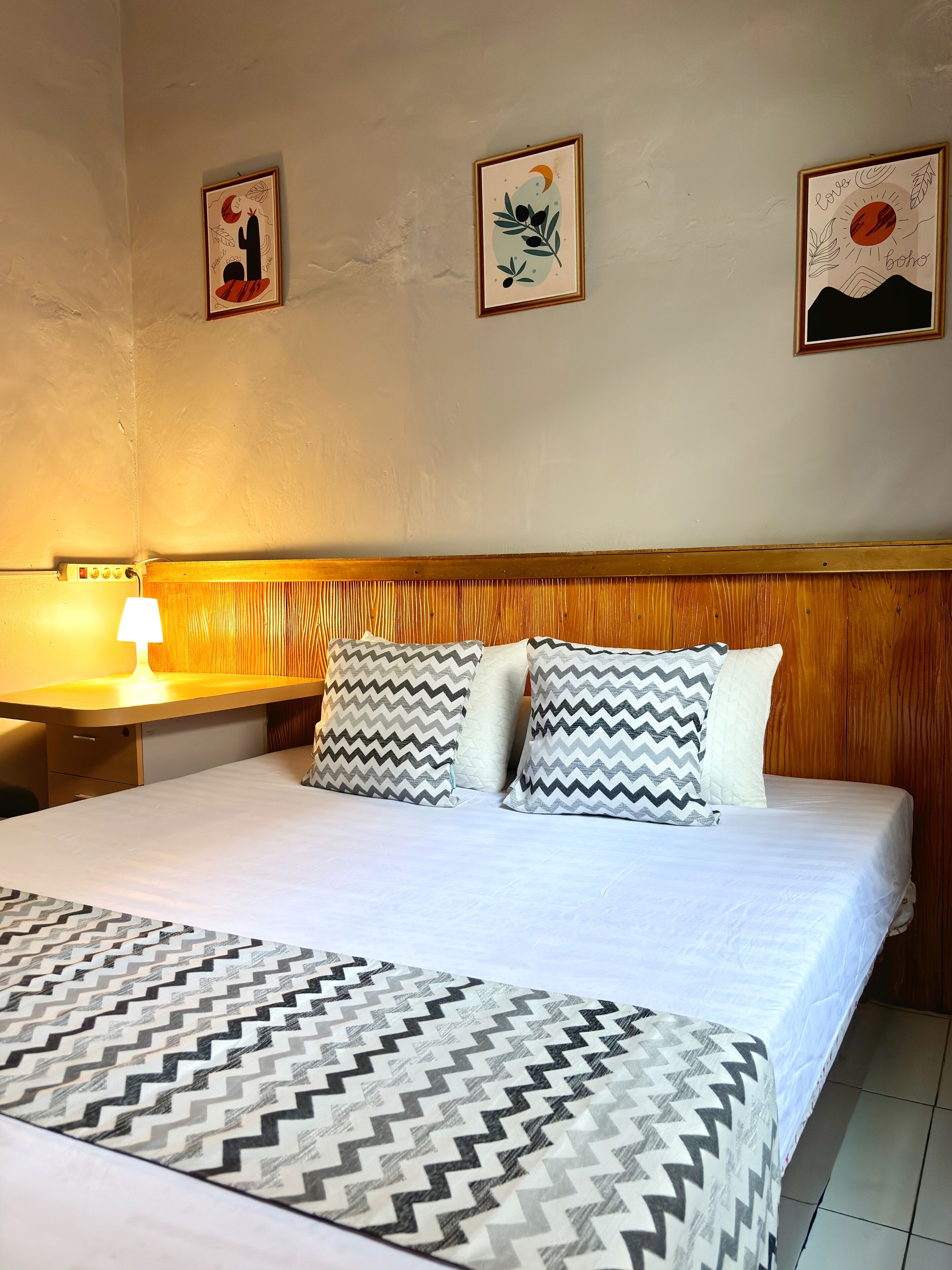 A hotel room with a wooden headboard, a bed with a patterned bedspread, two pillows with zigzag patterns, and three framed artworks on the wall.