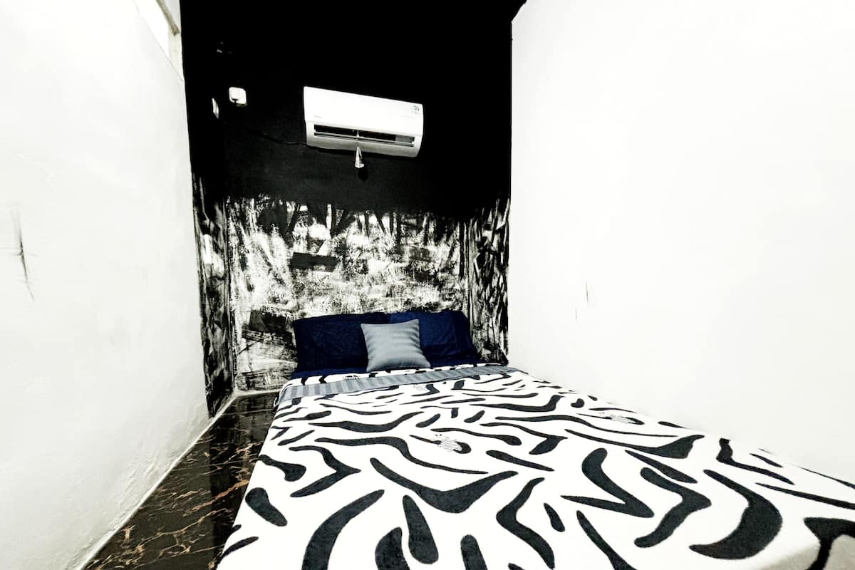A narrow room with a bed featuring a zebra print bedspread, a white air conditioning unit on the wall, and a dark floor.