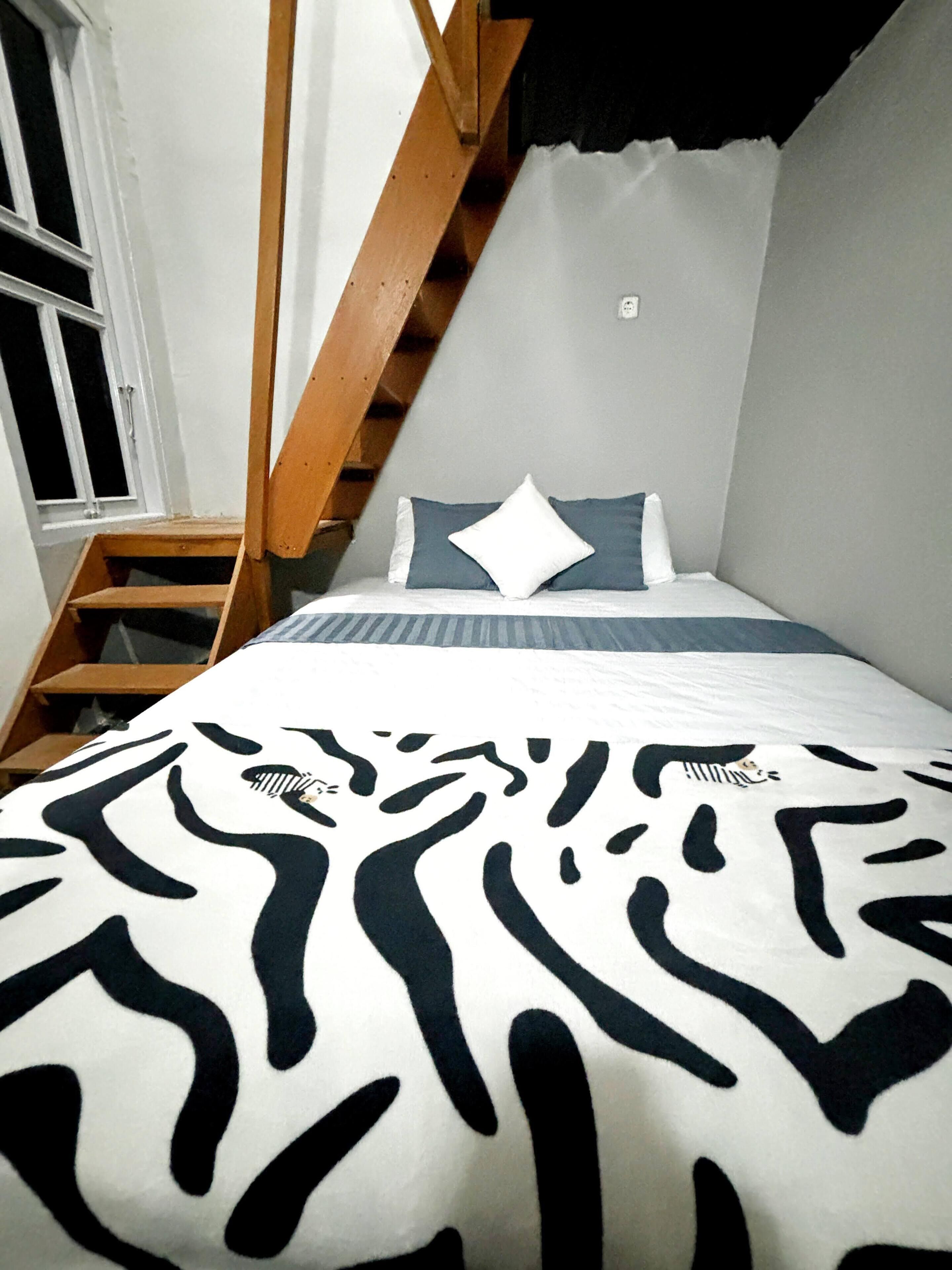 Een slaapkamer met een bed, een raam, een houten trap en een wandlamp.