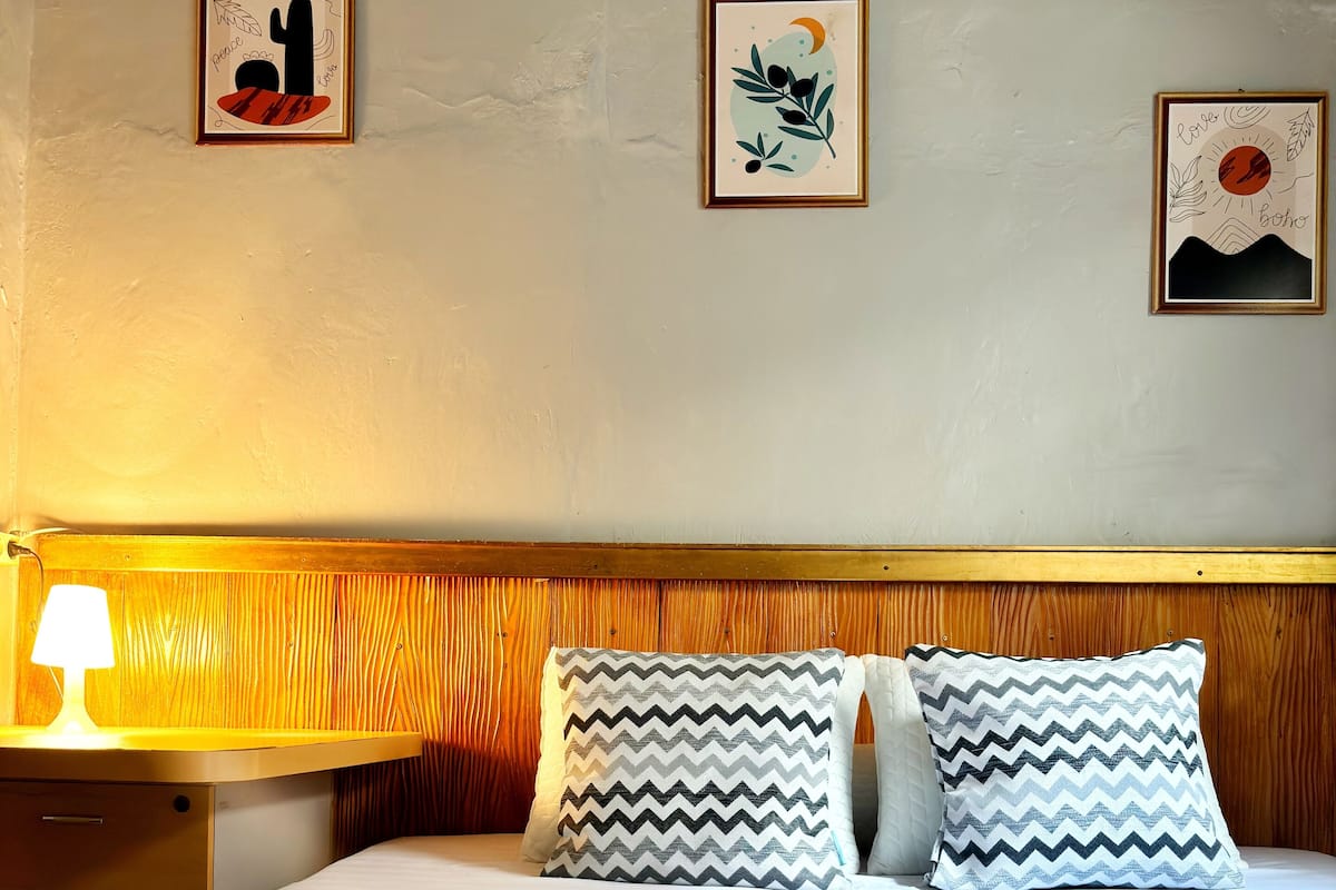 A hotel room with a bed, two pillows, a bedside table with a lamp, and three framed artworks on the wall.