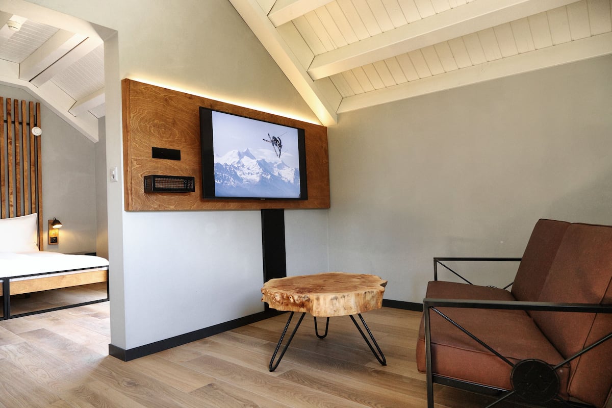 Une chambre moderne avec un plancher en bois, un téléviseur fixé au mur, une table d’appoint en bois et un fauteuil en cuir marron.