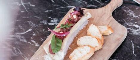 Ein Esstisch mit einem Holzservierbrett, das ein Sandwich, Brotscheiben, ein Weinglas und ein Wasserglas trägt.