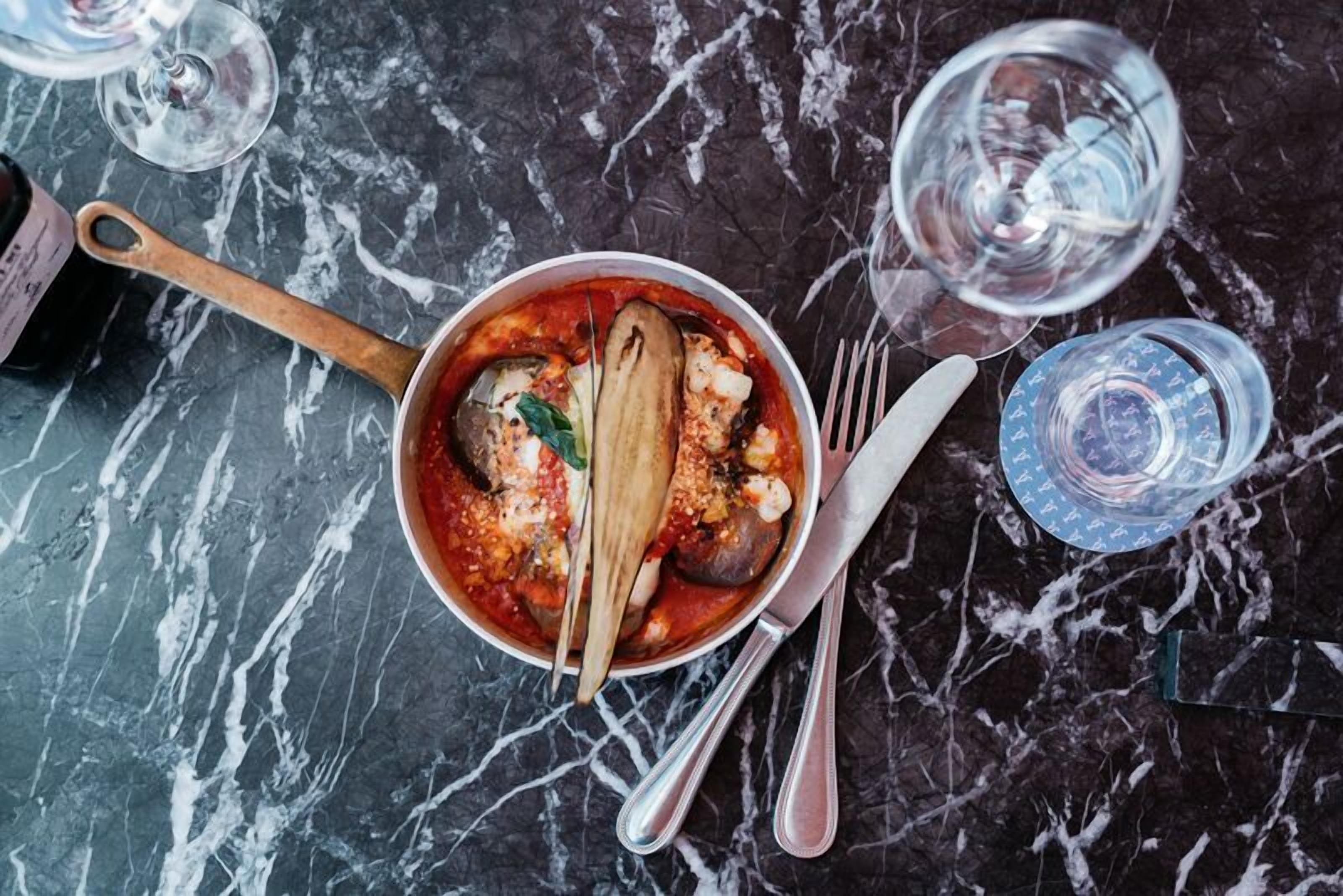 A bowl of food on a marble table with a spoon and fork, a bottle, and glasses.