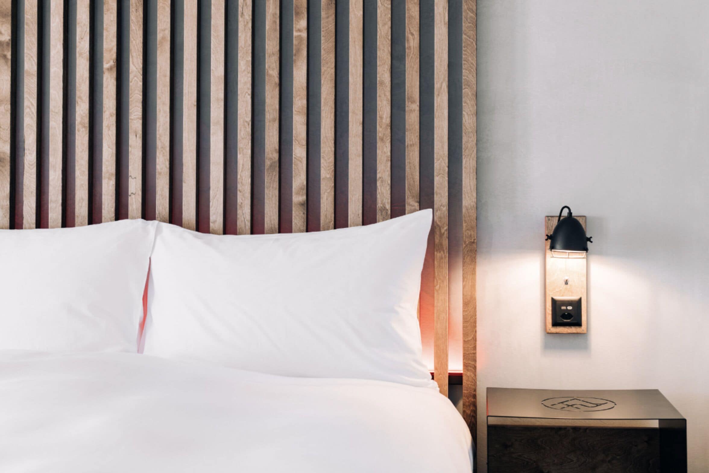 A neatly made bed with white linens, a wooden slat headboard, a wall-mounted nightstand with a lamp, and a small wooden box with a logo.