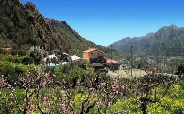A mountainous landscape with a building featuring a red roof, surrounded by lush greenery and flowering trees. - Agriturismo Ghiritina (Francavilla di Sicilia)