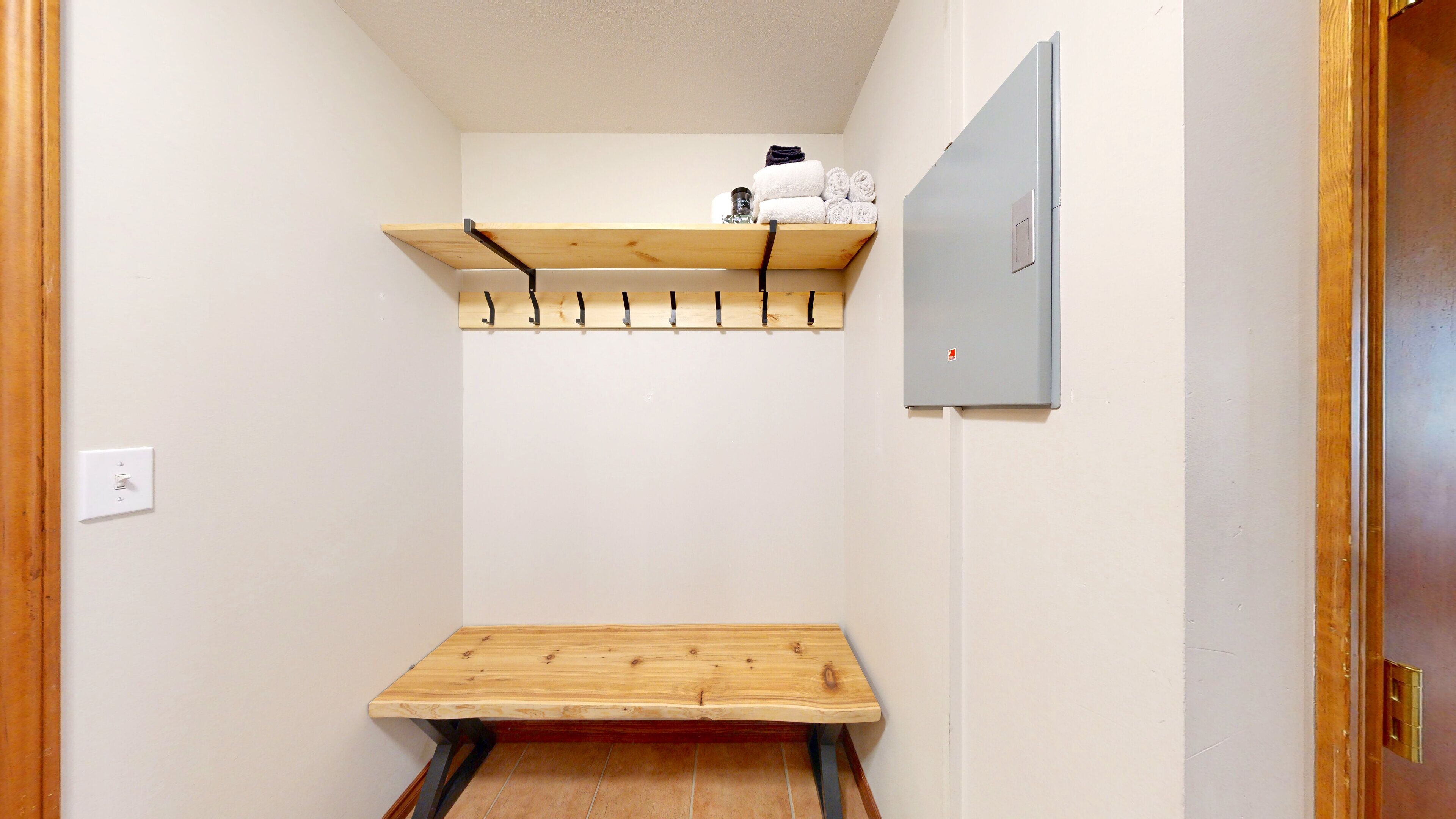 Un pasillo estrecho y pequeño con un banco de madera al final, un estante sobre él con toallas y un tarro de vidrio, y un enchufe en la pared izquierda.