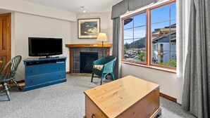 A living room with a television, blue dresser, green chair, wooden table, fireplace, and a large window with a view of buildings and hills.