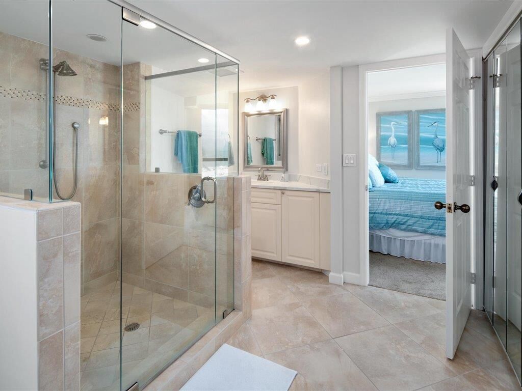 A modern bathroom with a glass-enclosed shower, a vanity with a mirror, and a view into a bedroom with a bed and artwork.