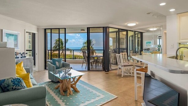 A modern living room with a large glass door leading to a balcony, a dining area, and a kitchen with a large TV. - Beachfront 2BR/2BA Sandcastle condo with pool, WiFi, and heated pool (Marco Island)