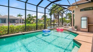 En swimmingpool med klart, blåt vand, omgivet af et glasindhegning, samt et loungeområde med stole og et lille bord.