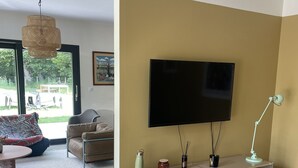 A modern living room with a flat-screen TV, a sofa, a coffee table, and a dining area with a table and chairs. - Maison d’architecte en bois (Chevry)
