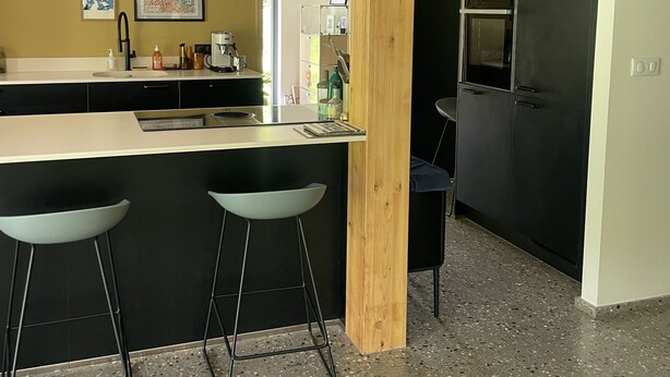 A modern kitchen with a wooden beam ceiling, a black countertop, and two bar stools. - Maison d’architecte en bois (Chevry)
