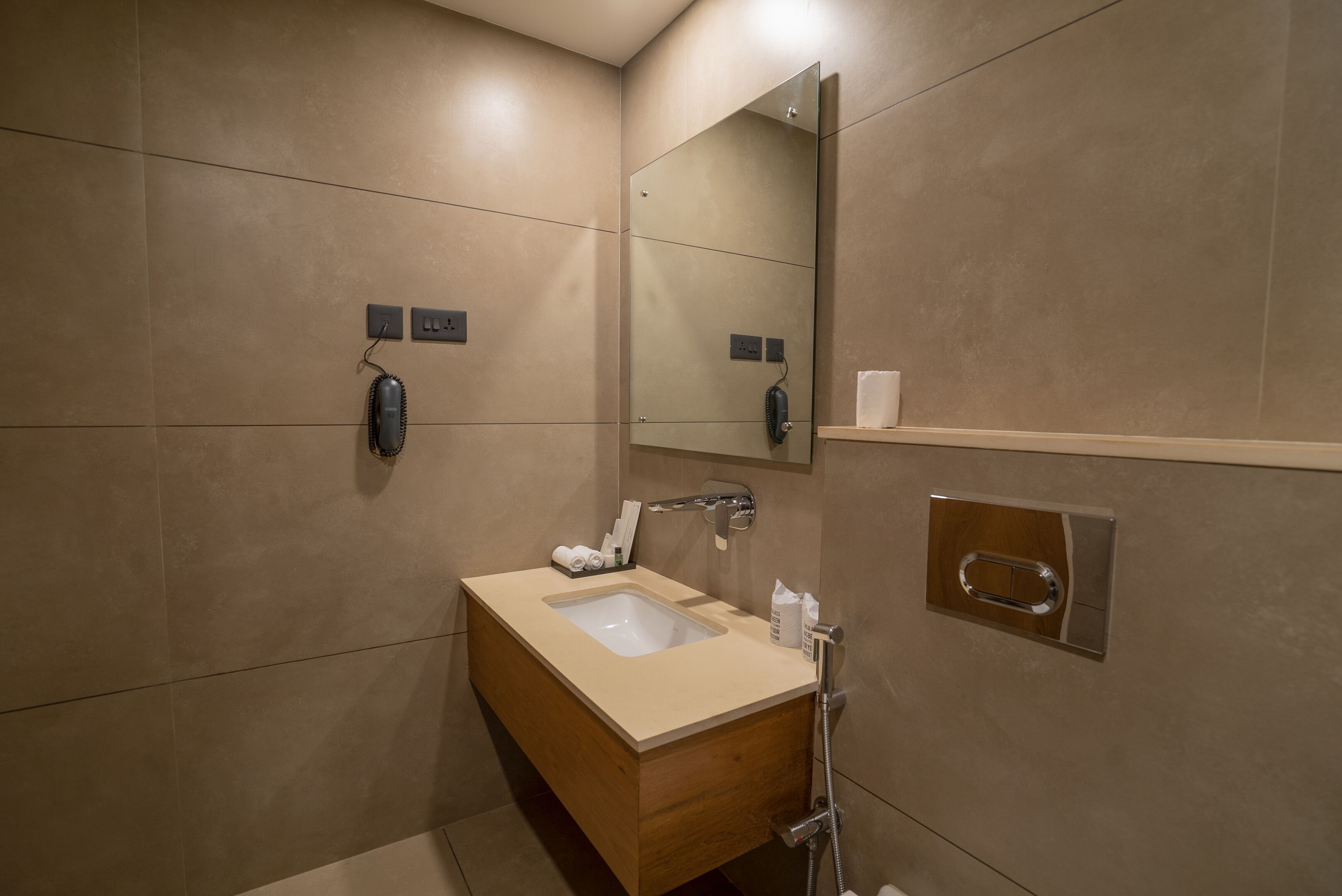 A modern bathroom with a wooden vanity, a large mirror, a wall-mounted telephone, and a shelf with toiletries.