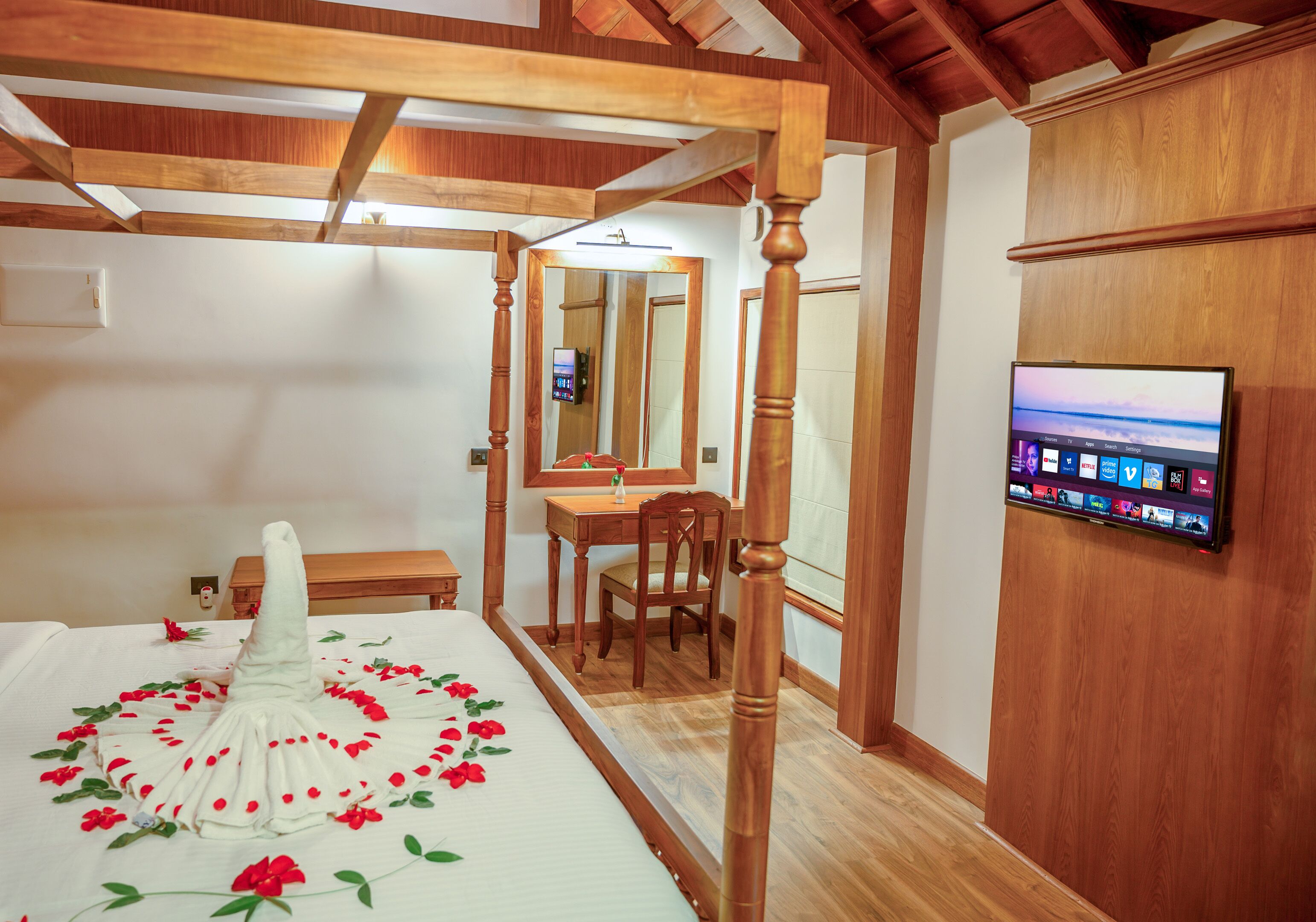 A bedroom with a four-poster bed, a wooden desk, a chair, a television mounted on the wall, and a mirror.
