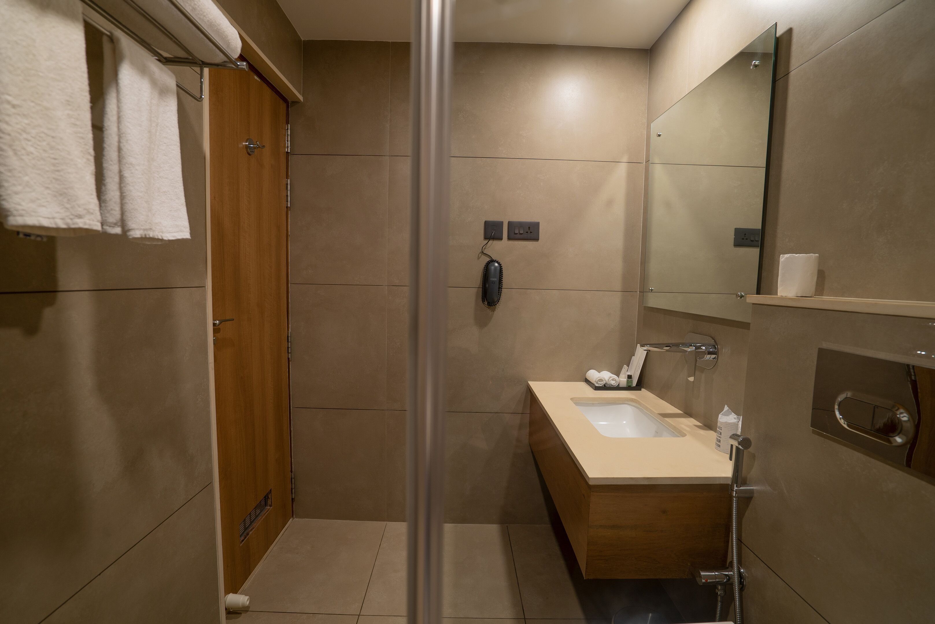 A modern bathroom with a wooden door, a glass shower enclosure, a beige sink, and a mirror.