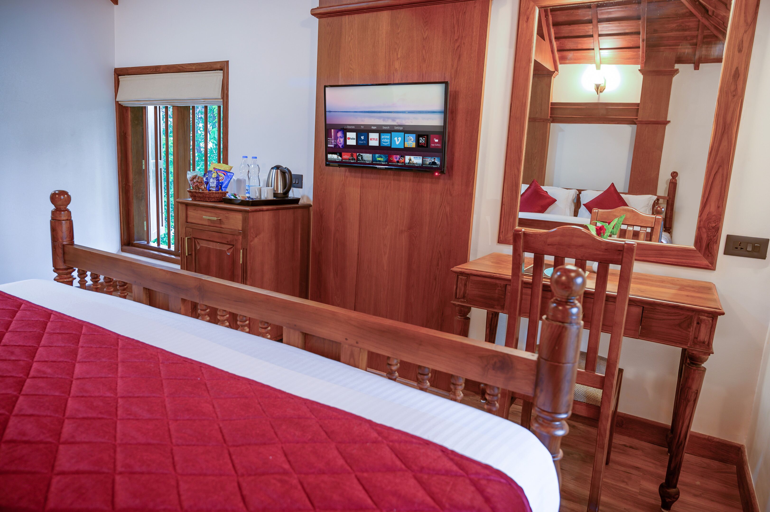 A hotel room with a wooden bed, a desk with a chair, a television mounted on the wall, and a window with a view of trees.