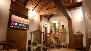 Un salón acogedor con techo de madera, un televisor de pantalla plana montado en la pared, una mesa de centro redonda con una planta y un comedor con mesa y sillas.