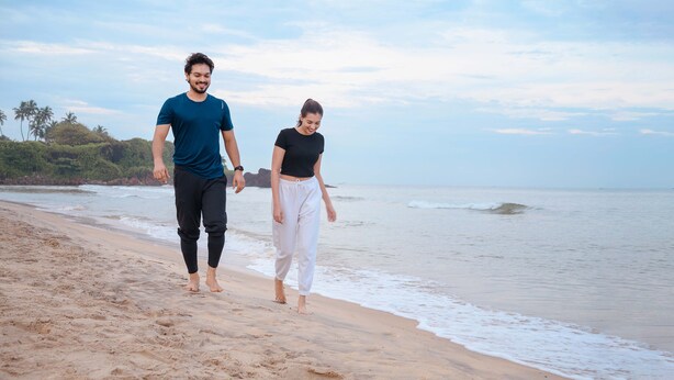 Una pareja caminando sobre una playa arenosa con un mar tranquilo y follaje al fondo.