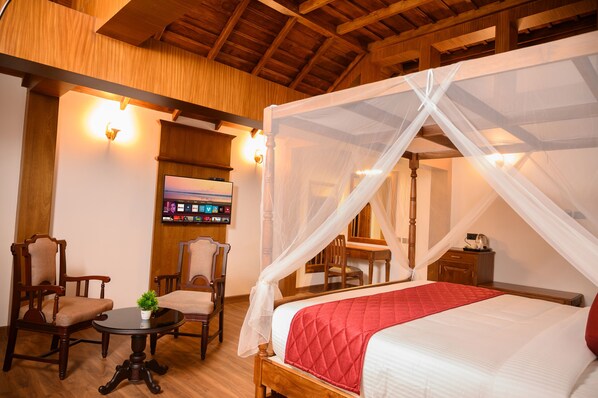 A room with a four-poster bed, wooden furniture, a small table with a plant, and a television on the wall. - Surya Heritage Hotels (Kannur)