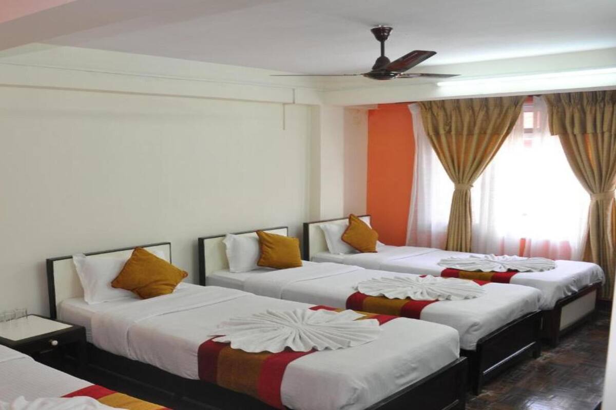 A hotel room with four single beds, each with white bedding and decorative pillows. There is a ceiling fan, a window with curtains, and a wooden floor.