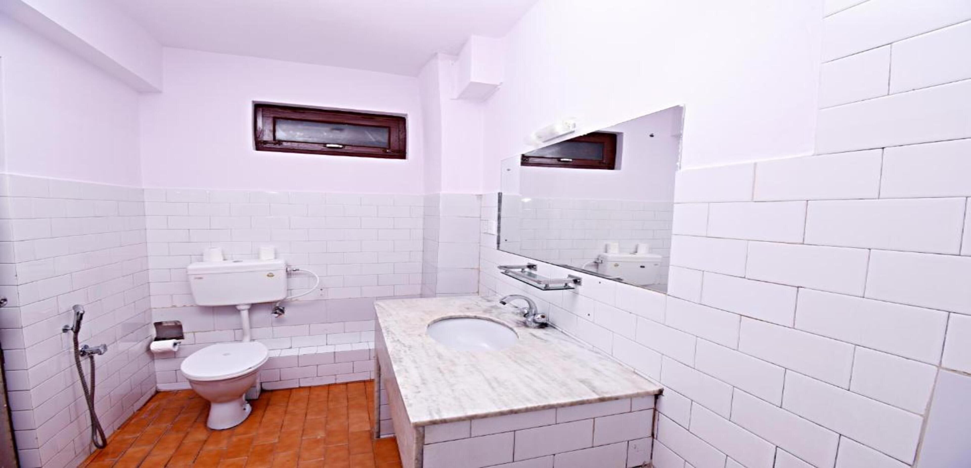 A small bathroom with white tiled walls, a white toilet, a sink with a mirror, and a window above the toilet.