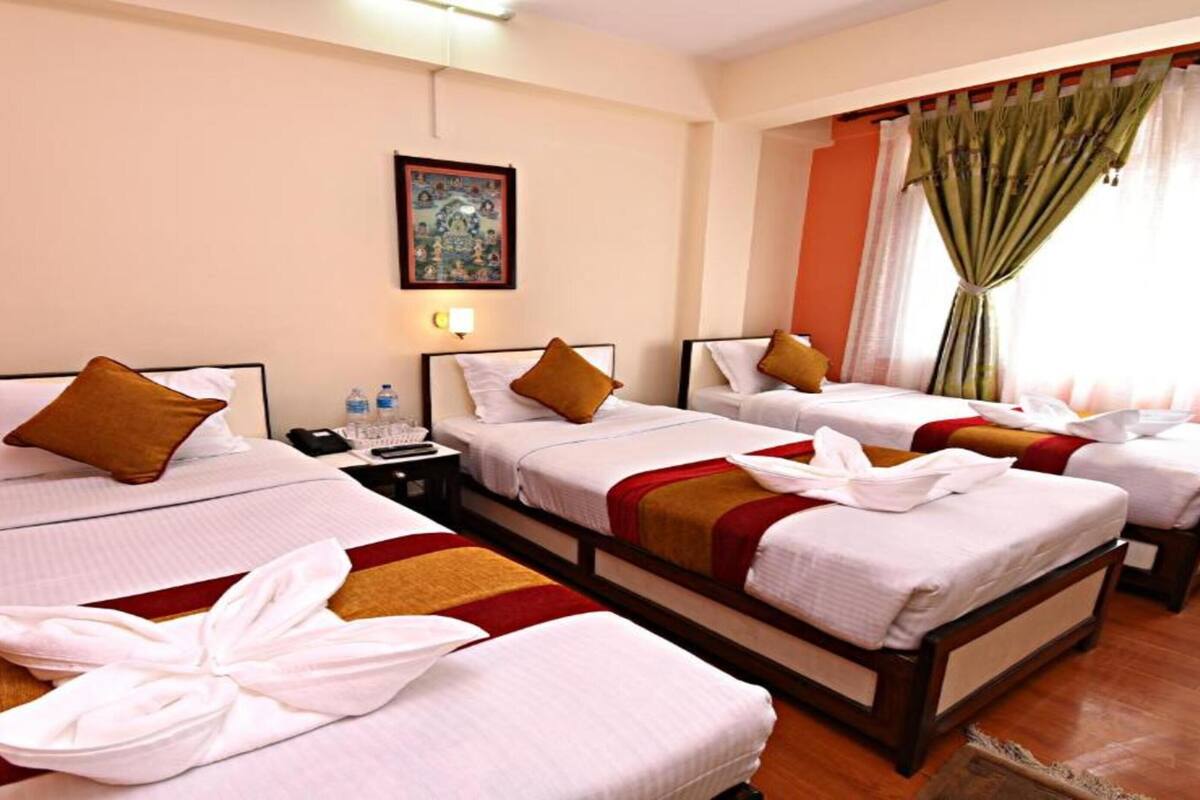 A hotel room with three single beds, each with a pillow and a neatly folded towel. There is a small table with a phone and a water bottle. The room has a painting on the wall and green curtains on the window.