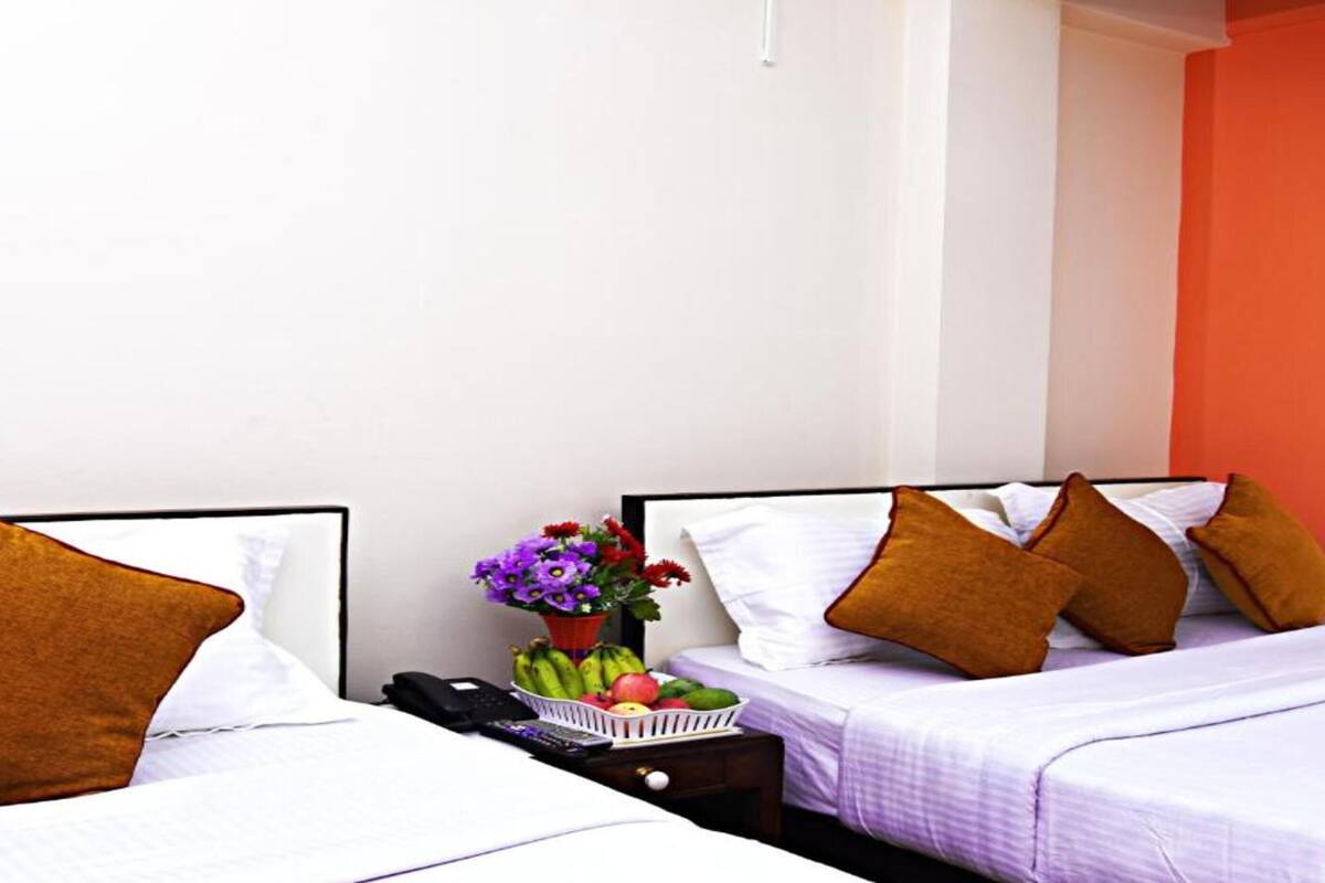 A hotel room with two beds, each with white bedding and brown pillows. There is a nightstand with a phone, a basket of fruit, and a vase of flowers.
